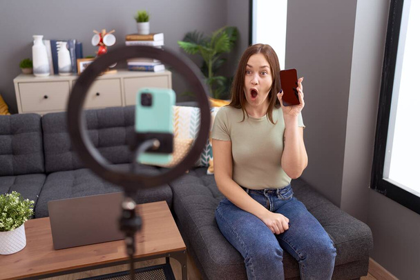 Beautiful brunette woman recording vlog tutorial with smartphone at home scared and amazed with open mouth for surprise, disbelief face  - Photo, Image