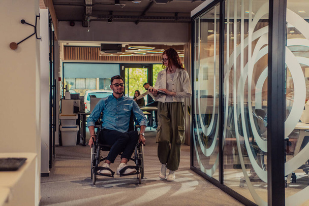 Young business colleagues, collaborative business colleagues, including a person in a wheelchair, walk past a modern glass office corridor, illustrating diversity, teamwork and empowerment in the - 写真・画像
