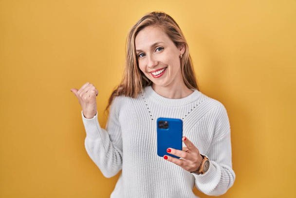 Giovane donna bionda utilizzando smartphone digitando messaggio che punta alla schiena dietro con mano e pollici in su, sorridendo fiducioso  - Foto, immagini