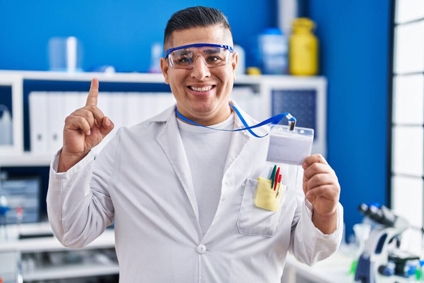 Joven hispano trabajando en laboratorio científico con tarjeta de identificación sonriendo con una idea o pregunta apuntando con el dedo con la cara feliz, número uno  - Foto, imagen