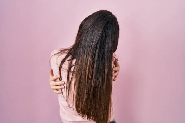 Young brunette woman standing over pink background hugging oneself happy and positive from backwards. self love and self care  - 写真・画像