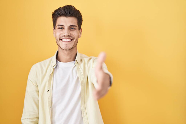 Молодой латиноамериканец, стоящий на желтом фоне, улыбаясь, дружелюбно предлагая рукопожатие в качестве приветствия и приветствия. успешный бизнес.  - Фото, изображение