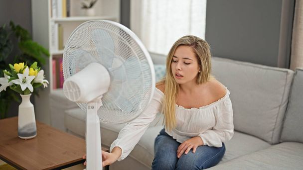 jong blond vrouw zitten op bank lijden voor Hot start ventilator thuis - Foto, afbeelding