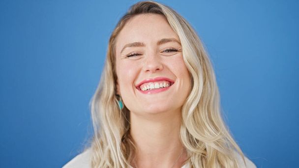 Young blonde woman smiling confident standing over isolated blue background - 写真・画像