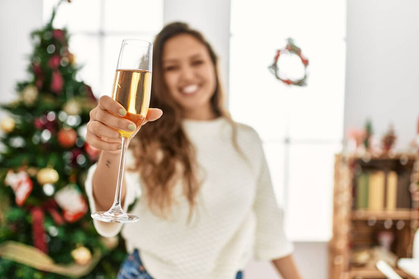jong mooi latino vrouw drinken champagne stand by kerstboom thuis - Foto, afbeelding