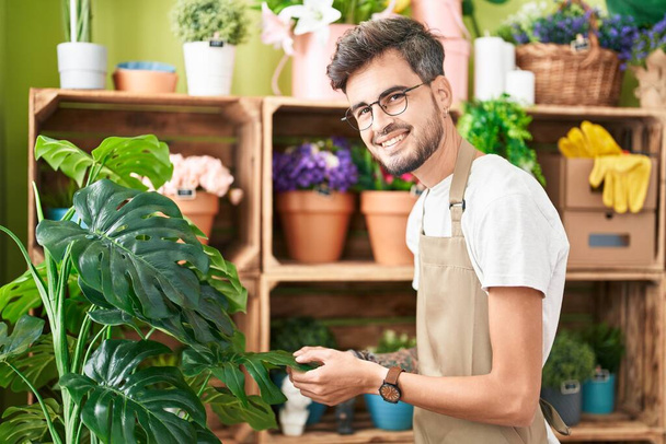 Junger hispanischer Florist lächelt selbstbewusst und hält Blechpflanze im Blumenladen - Foto, Bild