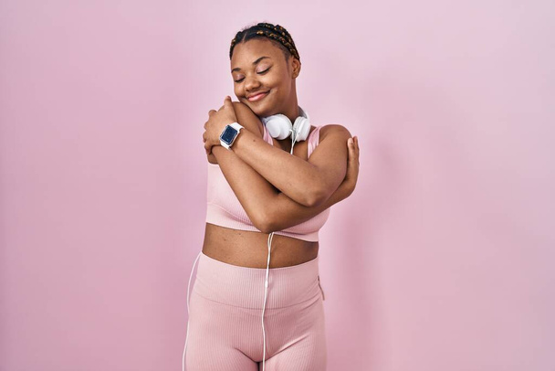 Mujer afroamericana con trenzas que usan ropa deportiva y auriculares abrazándose feliz y positivo, sonriendo confiado. amor propio y cuidado personal  - Foto, imagen