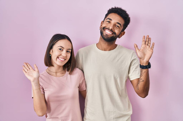 Молодая латиноамериканская пара вместе за розовым фоном отказ сказать привет счастливый и улыбающийся, дружественный жест приветствия  - Фото, изображение