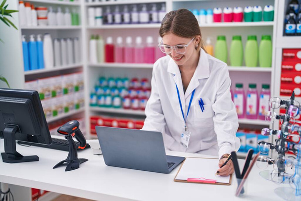 Молодая блондинка фармацевт с помощью ноутбука записи на документе в аптеке - Фото, изображение
