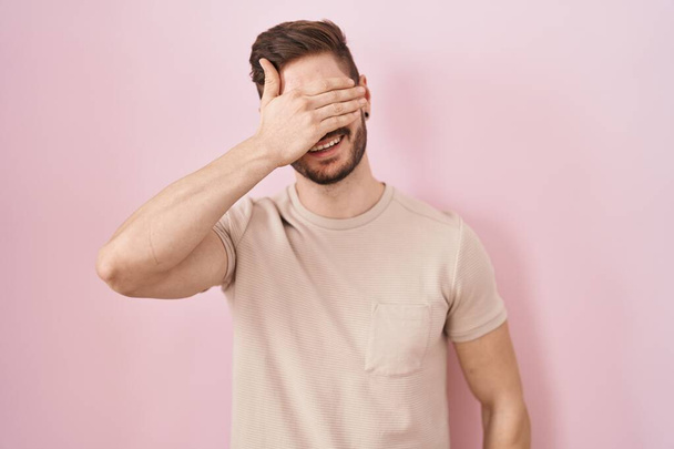 Латиноамериканець з бородою стоїть над рожевим фоном, усміхаючись і сміючись рукою на обличчі, закриваючи очі на подив. сліпа концепція.  - Фото, зображення