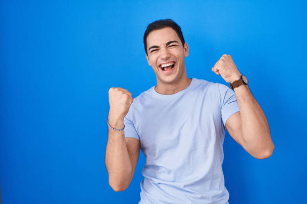 Young hispanic man standing over blue background celebrating surprised and amazed for success with arms raised and eyes closed. winner concept.  - Foto, Imagen