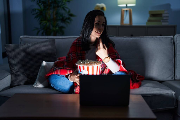 Hispanic woman eating popcorn watching a movie on the sofa beckoning come here gesture with hand inviting welcoming happy and smiling  - Photo, Image