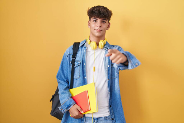 Hispánský teenager v studentském batohu a s knihami ukazujícími prstem na kameru se šťastnou a zábavnou tváří. dobrá energie a vibrace.  - Fotografie, Obrázek