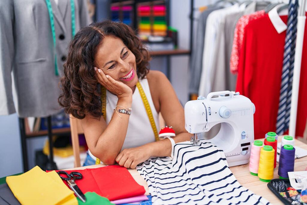 Femme d'âge moyen tailleur souriant confiant assis sur la table à l'usine de vêtements - Photo, image