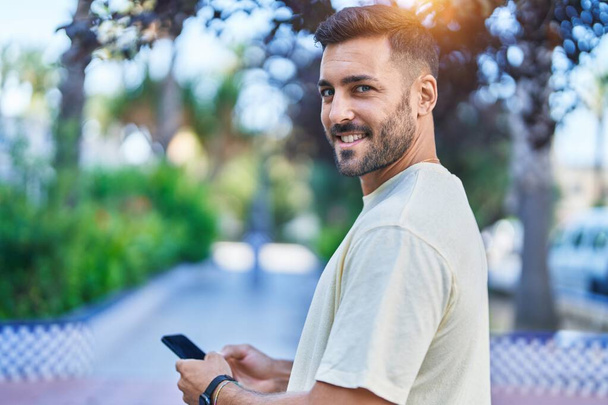 Giovane uomo ispanico sorridente fiducioso utilizzando smartphone al parco - Foto, immagini