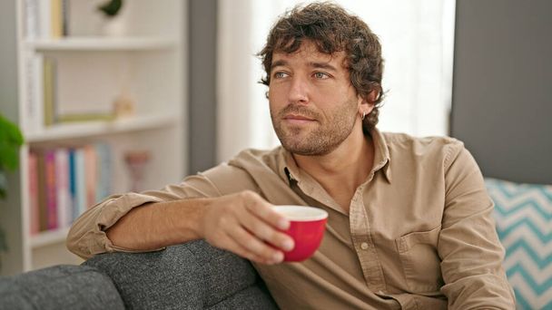 Young hispanic man holding cup of coffee sitting on sofa at home - Foto, afbeelding