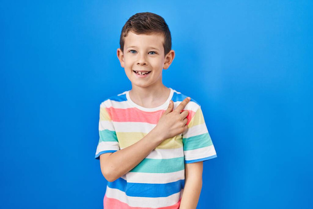 Молодой кавказский ребенок стоит на синем фоне веселый с улыбкой на лице указывая рукой и пальцем в сторону со счастливым и естественным выражением  - Фото, изображение