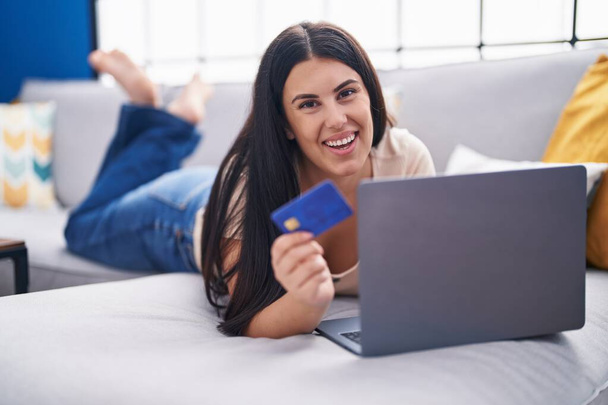 Joven mujer hispana hermosa usando laptop y tarjeta de crédito tumbado en el sofá en casa - Foto, Imagen