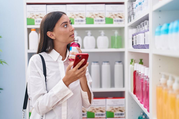 Young beautiful hispanic woman customer using smartphone holding medicine bottle at pharmacy - Photo, Image