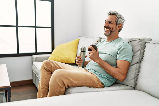 Grauhaariger Mann mittleren Alters trinkt Mate-Infusion zu Hause auf Sofa - Foto, Bild