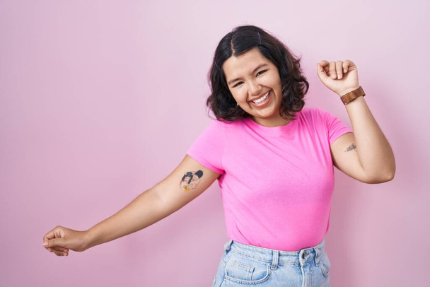 ピンクの背景の上に立つ若いヒスパニック系の女性ダンス幸せと陽気です,笑顔移動カジュアルで自信を持って音楽に耳を傾け  - 写真・画像