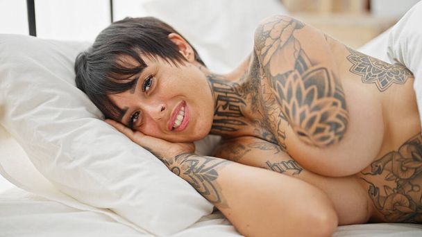 Hispanische Frau mit amputiertem Arm liegt auf Bett und lächelt hemdlos ins Schlafzimmer - Foto, Bild