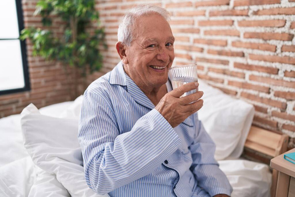 Mężczyzna w średnim wieku, siwe włosy, pijący szklankę wody, siedzący na łóżku w sypialni. - Zdjęcie, obraz