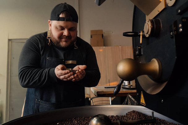 Trabajador masculino en gorra y uniforme negro de pie cerca de la máquina de asar con un puñado de granos de café y granos tostados que huelen - Foto, Imagen
