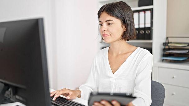 Young beautiful hispanic woman business worker using touchpad and computer at office - Photo, Image