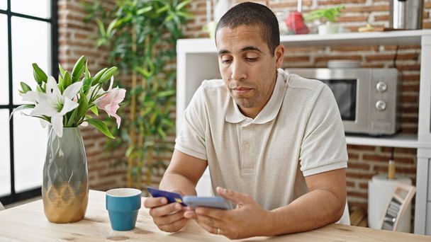 Jeune homme hispanique faisant du shopping avec smartphone et carte de crédit assis sur la table à manger - Photo, image
