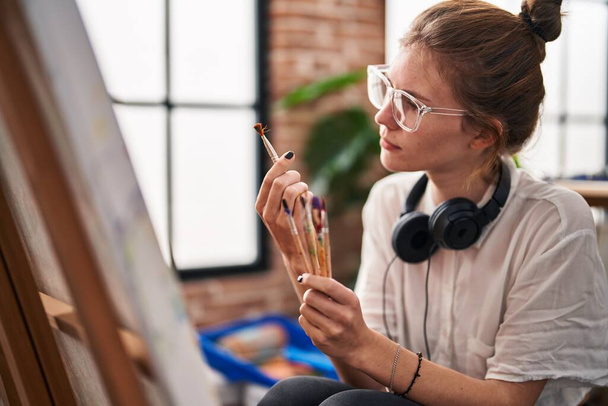 アートスタジオで絵筆を見ている若い金髪の女性アーティスト - 写真・画像