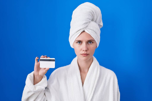 Mulher branca loira vestindo roupão segurando cartão de crédito pensando atitude e expressão sóbria olhando auto confiante  - Foto, Imagem