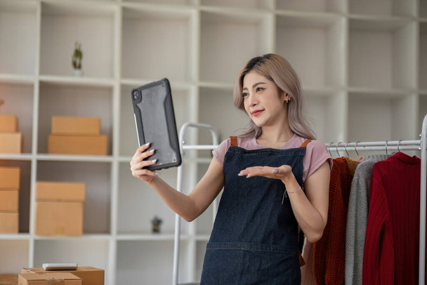 businesswoman owner an online trading business is using tablet to check the stock of the clothes on the rail rack at small office at home. work from home, online delivery, SME.. - Photo, image