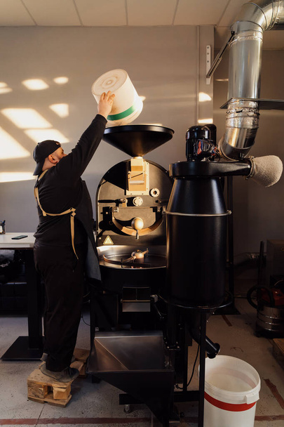 Boční pohled mužského pracovníka v uniformě a čepici nalévání syrových kávových zrn do zásobníku profesionálního pražicího stroje při práci v továrně - Fotografie, Obrázek
