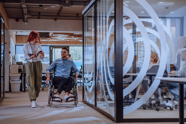 Young business colleagues, collaborative business colleagues, including a person in a wheelchair, walk past a modern glass office corridor, illustrating diversity, teamwork and empowerment in the - Foto, Imagem