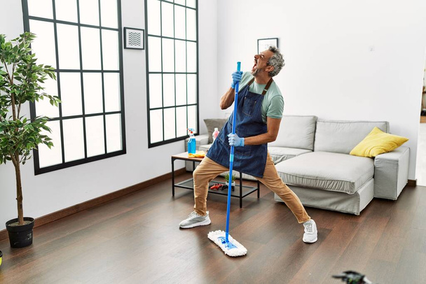 Middle age grey-haired man singing song using mop as a microphone at home - Photo, Image