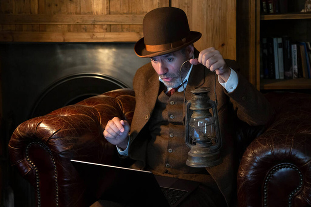 Een man met een hoed en een Victoriaans kostuum met een antieke lamp in zijn hand kijkt in retrostijl naar de laptop. Het thema is moderne technologie en de vorige eeuw. - Foto, afbeelding