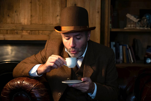 Un homme en costume et chapeau victorien boit le café d'une tasse en porcelaine blanche dans un salon de style rétro. - Photo, image