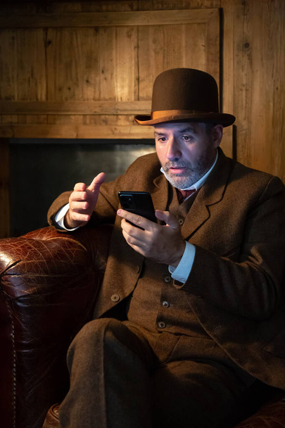 Un hombre con sombrero y un disfraz victoriano mira el teléfono móvil con sorpresa en un salón de estilo retro. El tema es la tecnología moderna y el siglo pasado. Imagen vertical. - Foto, Imagen
