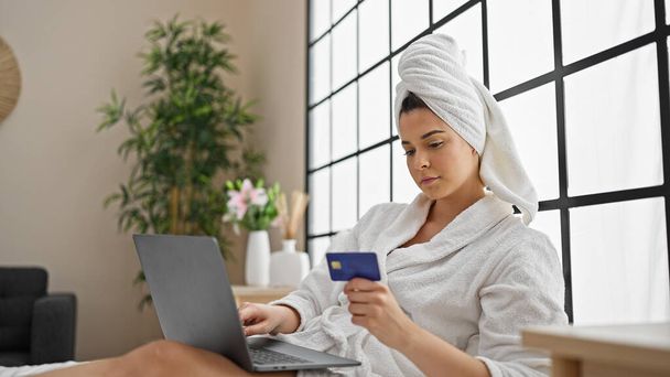 Young beautiful hispanic woman wearing bathrobe shopping with laptop and credit card at bedroom - Фото, изображение