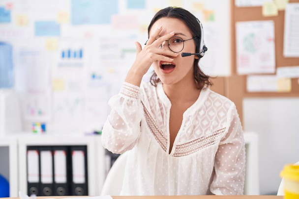 Latynoska młoda kobieta pracująca w biurze w słuchawkach i okularach zaglądająca ręką w szok zakrywający twarz i oczy, patrząca przez palce przestraszona  - Zdjęcie, obraz