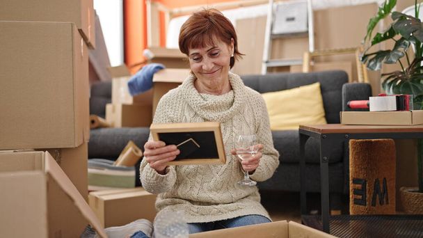 Mujer hispana madura desempacando caja de cartón bebiendo vino en un nuevo hogar - Foto, imagen
