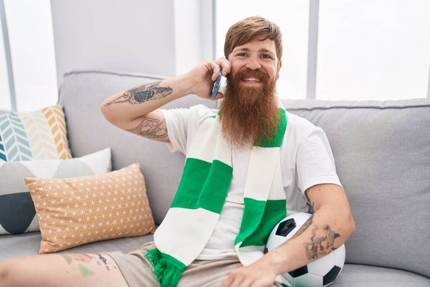自宅でスマートフォンで話すサッカーチームをサポートする若い赤毛の男 - 写真・画像