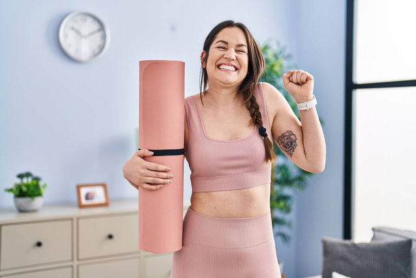 jong brunette vrouw holding yoga mat schreeuwen trots, vieren overwinning en succes zeer enthousiast met opgeheven arm  - Foto, afbeelding