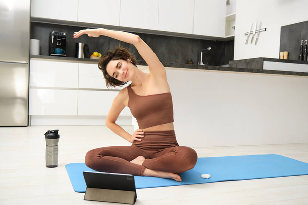 Dijital tablete bakan genç bir kadın, internetteki spor salonuna bağlanıyor, antrenman yapıyor, evdeki yoga paspasına oturuyor, beden eğitimi egzersizlerini uzaktan takip ediyor.. - Fotoğraf, Görsel