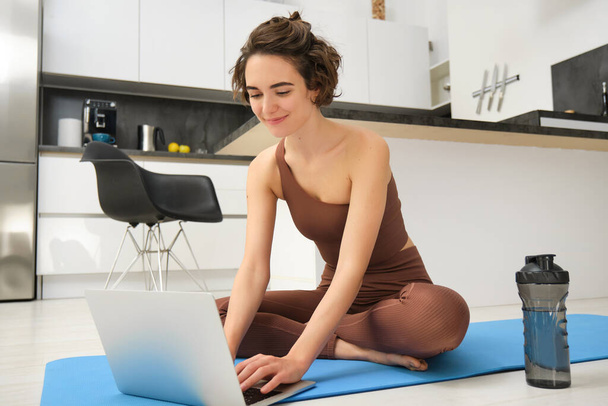 Портрет молодой спортсменки, инструктора по фитнесу, начинающей заниматься онлайн гимнастикой у себя дома, сидящей на резиновой коврике для йоги, печатающей на ноутбуке. Спорт и стиль жизни - Фото, изображение