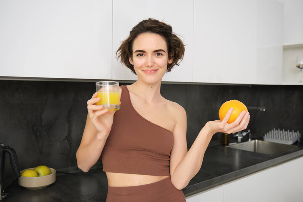Estilo de vida saudável e esporte. Mulher sorridente bonita, bebendo suco de laranja fresco e segurando frutas na mão, desfrutando de sua bebida vitamínica após o treino em casa, de pé na cozinha. - Foto, Imagem