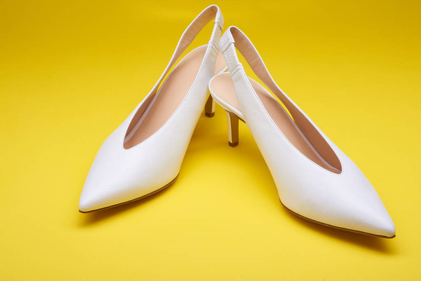 Αριστοκρατικά λευκά γυναικεία δερμάτινα ανοιχτόχρωμα ψηλοτάκουνα παπούτσια με πίσω τιράντες, απομονωμένα σε φωτεινό κίτρινο φόντο. Κοντινή θέα. Μοντέρνα γυναικεία παπούτσια, παπούτσια για τη νύφη - Φωτογραφία, εικόνα