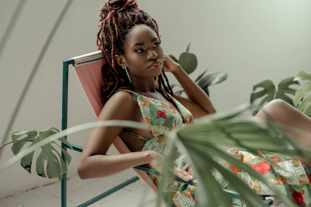 Mulher africana encantadora, vestindo um vestido de ornamento multicolorido, sentada em uma cadeira de rede cercada por plantas tropicais. Descansando em atmosfera calmante. Bem-estar, bem-estar, conceito de atenção plena. - Foto, Imagem