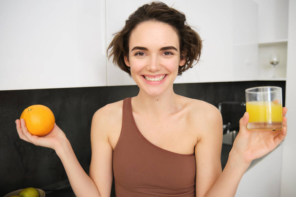 Gülümseyen sporcu kadın portakal suyu ve bir bardak meyve suyu gösteriyor, sağlıklı ve aktif yaşam tarzının ipuçlarını paylaşıyor, mutfakta ayakta duruyor.. - Fotoğraf, Görsel